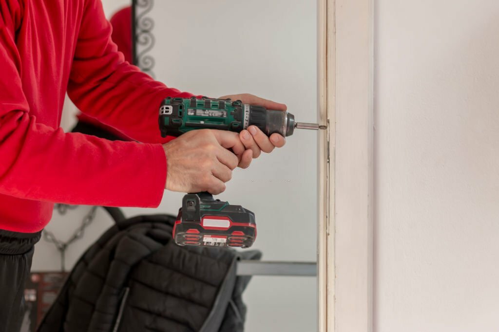 Mature male locksmith installing a lock on an old white door with a hand drill